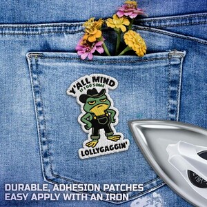 May include: Embroidered patch of a green frog in a cowboy hat and overalls, with the text "Y'all Mind If I Do Some Lollygaggin'". The patch is on denim, with a pocket holding flowers. An iron is in the foreground.
