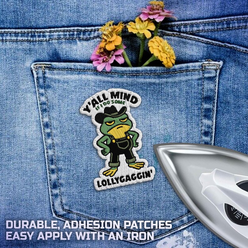 May include: Embroidered patch of a green frog in a cowboy hat and overalls, with the text "Y'all Mind If I Do Some Lollygaggin'". The patch is on denim, with a pocket holding flowers. An iron is in the foreground.
