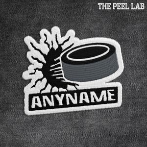 May include: Embroidered patch with a hockey puck and a stylized design. The patch has the text "ANYNAME" in a rectangular border. The text "THE PEEL LAB" is at the top. The design is in black, white, and grey.