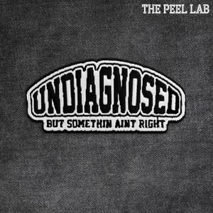 May include: A black and white embroidered patch with the text "UNDIAGNOSED" in a bold, stylized font. Below, the phrase "BUT SOMETHIN AINT RIGHT" is displayed. The patch is set against a textured gray background.