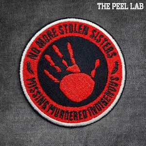 May include: Circular embroidered patch with the text "NO MORE STOLEN SISTERS" and "MISSING MURDERED INDIGENOUS" in red on a white border. A red handprint is centered on a navy blue background. The text "THE PEEL LAB" is in the upper right corner.
