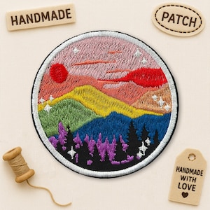 May include: Embroidered round patch featuring a colorful mountain landscape with a red sun and pink sky. The patch has a black border and the words "HANDMADE" and "PATCH" are on separate wooden tags. A wooden tag reads "HANDMADE WITH LOVE".