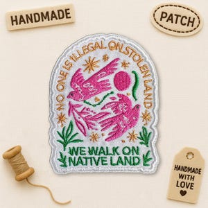 May include: Embroidered patch featuring pink birds and the text "NO ONE IS ILLEGAL ON STOLEN LAND" and "WE WALK ON NATIVE LAND." The patch has white, green, and gold detailing. Includes wooden tags that say "HANDMADE" and "HANDMADE WITH LOVE."