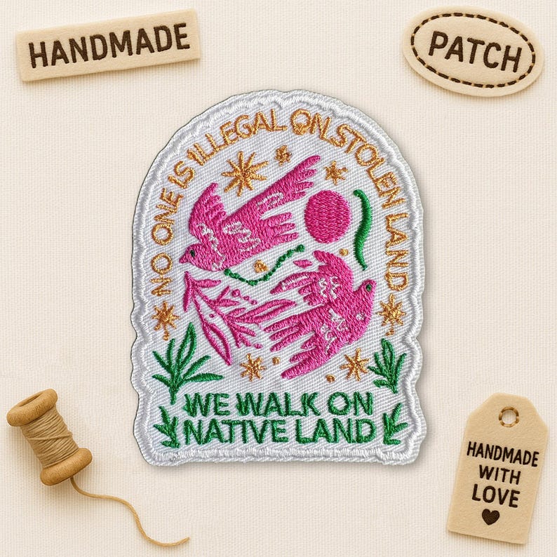 May include: Embroidered patch with pink birds, text reading "NO ONE IS ILLEGAL ON STOLEN LAND" and "WE WALK ON NATIVE LAND." The patch is white with green and gold accents. Includes wooden tags that say "HANDMADE" and "HANDMADE WITH LOVE."