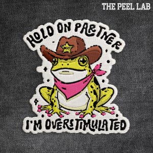 May include: Embroidered patch featuring a cartoon frog wearing a cowboy hat and pink bandana. Text reads "HOLD ON PARTNER I'M OVERSTIMULATED" in white lettering. The frog is yellow with black spots and a brown hat.