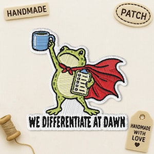 May include: An embroidered patch featuring a green frog wearing a red cape, holding a blue mug and a clipboard. Below the frog, the text reads WE DIFFERENTIATE AT DAWN. The patch is surrounded by a handmade label and a patch label on a light fabric.