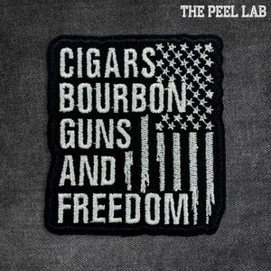 May include: Black and white embroidered patch with the words "CIGARS BOURBON GUNS AND FREEDOM" arranged vertically. The design incorporates an American flag motif with stars and stripes. The Peel Lab logo is in the top right corner.