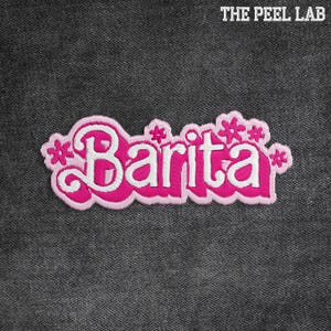 May include: An embroidered patch in pink and white, featuring the name "Barita" in a decorative font. The patch has a pink border and floral accents. The text "THE PEEL LAB" is at the top.