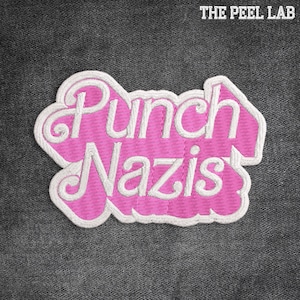 May include: A pink embroidered patch with the words "Punch Nazis" in a stylized font, outlined in white. The patch is set against a dark grey textured background, with the text "THE PEEL LAB" at the top.