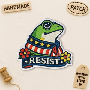 May include: Embroidered patch featuring a green frog wearing a patriotic scarf with the word "RESIST". The patch includes red, white, and blue colors, with yellow stars and flowers. The word "HANDMADE" is on a wooden tag.