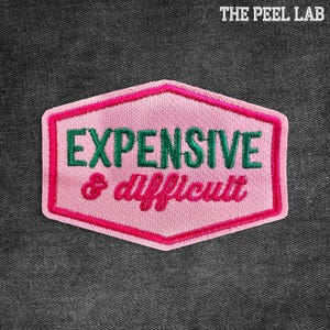 May include: A pink embroidered patch with the words "EXPENSIVE & difficult" in green and pink thread. The patch has a hexagonal shape with a pink border and is set against a dark gray fabric background. The text "THE PEEL LAB" is at the top.