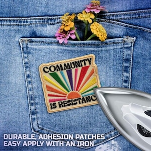 May include: A colorful patch with the words "COMMUNITY IS RESISTANCE" is ironed onto a blue denim pocket. The patch features a rainbow sun design. A few flowers are tucked into the pocket. An iron is in the lower right corner.
