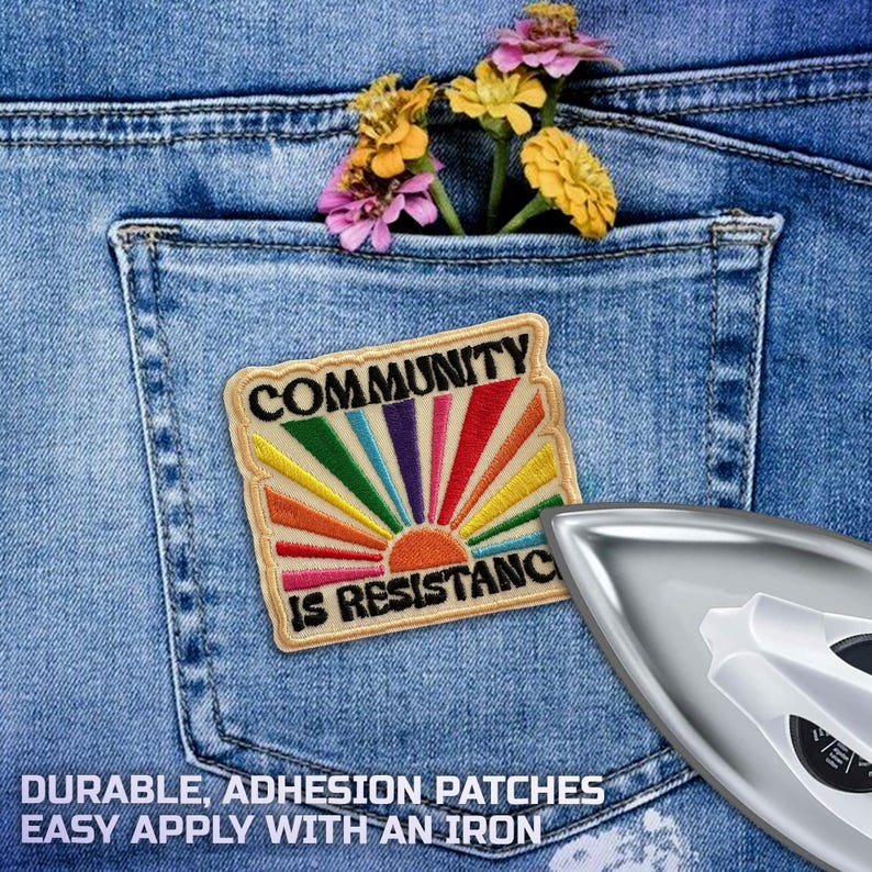 May include: A colorful patch with the words "COMMUNITY IS RESISTANCE" is ironed onto a blue denim pocket. The patch features a rainbow sun design. A few flowers are tucked into the pocket. An iron is in the lower right corner.