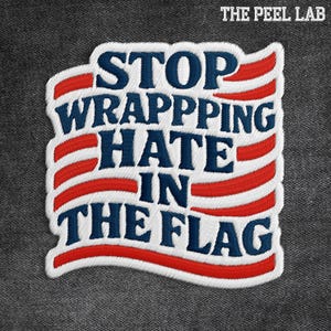 May include: Embroidered patch with the text "STOP WRAPPING HATE IN THE FLAG" in blue and red, resembling the American flag. The patch has a white border and is set against a gray fabric background.