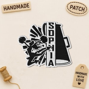 May include: Black and white embroidered patch featuring a cheerleader with pom-poms and the name "SOPHIA". The patch is in the shape of a megaphone. Additional tags read "HANDMADE" and "PATCH".