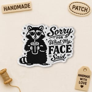May include: Embroidered patch featuring a raccoon wearing sunglasses and holding a drink, with the text "Sorry for What My Face Said." The patch is black and white with a white border. Includes a "Handmade" wooden tag and a "Patch" tag.