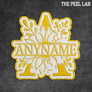 May include: Embroidered patch with a floral design and the word "ANYNAME" in gold thread. The patch features a yellow border and a white floral pattern. The text "THE PEEL LAB" is in the top right corner.