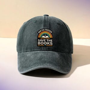 May include: A dark teal baseball cap with a curved brim. The cap features a patch with the text "BAN THE FASCISTS" above a rainbow and an open book, with "SAVE THE BOOKS" below. The cap is made of a soft, worn fabric.