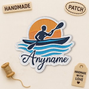 May include: Embroidered patch showing a kayaker on blue waves beneath an orange sun. The patch includes the word "Anymame" in cursive. Additional tags read "Handmade" and "Patch". A small tag says "Handmade with Love".