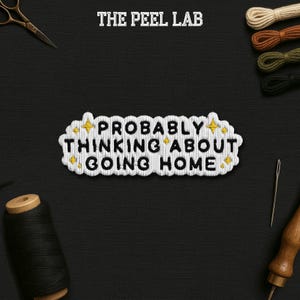 May include: Embroidered patch with the text "PROBABLY THINKING ABOUT GOING HOME" in black and white. The patch has a white border with yellow star accents. The words are stitched on a black background with sewing tools.