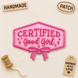 May include: A pink embroidered patch in a hexagonal shape with the words "CERTIFIED Good Girl" in a script font. The patch has a pink bow at the bottom and is surrounded by a pink border. The word "HANDMADE" is on a small wooden tag.