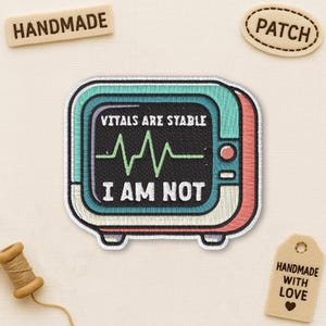 May include: Embroidered patch with a retro-style medical monitor design. The patch features the text "VITALS ARE STABLE" above a green EKG line, and "I AM NOT" below. The patch is bordered in teal, red, and cream colors.