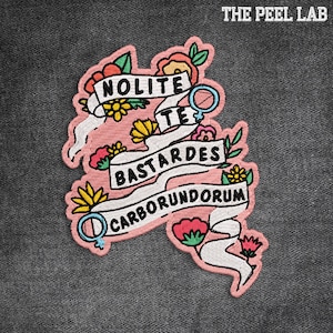 May include: Embroidered patch with a pink outline, featuring the text "Nolite Te Bastardes Carborundorum" on a white banner, surrounded by colorful flowers and leaves. The patch includes a female symbol and the text "THE PEEL LAB".