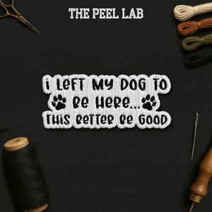 May include: Embroidered patch with the text "I LEFT MY DOG TO Be Here... THIS BETTER BE GOOD" in white thread. The patch includes paw prints and the text "THE PEEL LAB" at the top. The background is black, with sewing tools and thread spools.