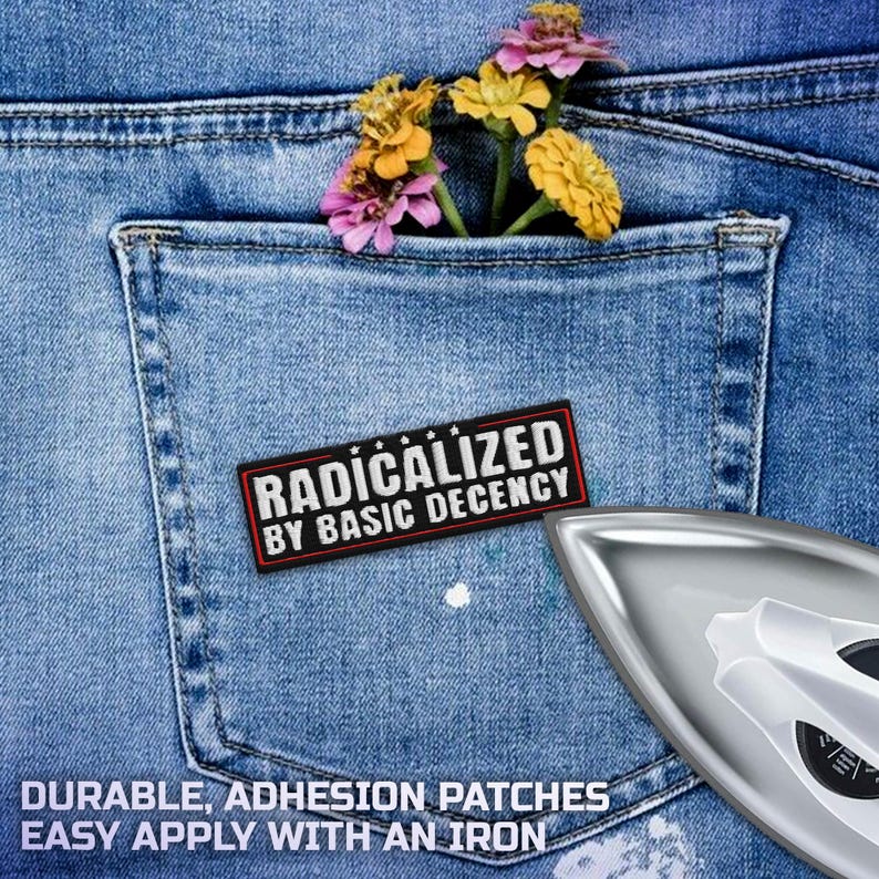 K&ouml;nnte beinhalten: Ein rechteckiger Aufn&auml;her mit dem Text "RADICALIZED BY BASIC DECENCY" in Wei&szlig; auf schwarzem Hintergrund mit rotem Rand, platziert auf einer blauen Jeans-Tasche. Die Tasche enth&auml;lt bunte Blumen. Ein B&uuml;geleisen ist im Bild.