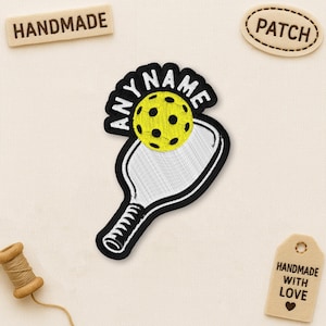 May include: Embroidered patch featuring a pickleball paddle and ball with the text "ANYNAME". The ball is yellow with black dots. The paddle is white with a black outline. Additional tags read "HANDMADE", "PATCH", and "HANDMADE WITH LOVE".