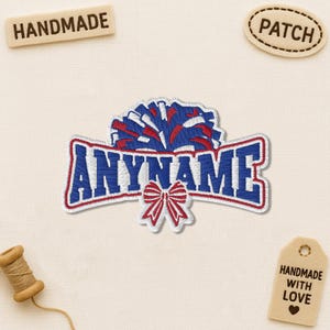 May include: Embroidered patch with the text "ANYNAME" in blue with a red outline, a red bow, and a pom-pom design in red, white, and blue. The patch is surrounded by a cream-colored background. Additional tags read "HANDMADE", "PATCH", and "HANDMADE WITH LOVE".