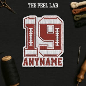 May include: Embroidered patch with the number 19 in red and white, with the text "ANYNAME" below. The patch is on a black background with sewing supplies, including scissors, thread, and a needle. The text "THE PEEL LAB" is at the top.
