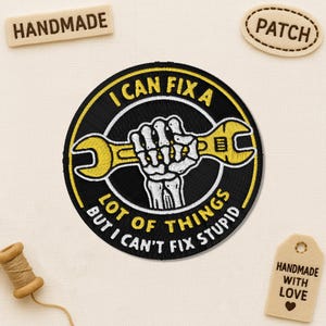 Peut inclure: Écusson brodé avec le texte "I CAN FIX A LOT OF THINGS BUT I CAN'T FIX STUPID" dans un design circulaire. L'écusson présente une clé anglaise jaune tenue par une main de squelette. Des étiquettes supplémentaires indiquent "HANDMADE" et "PATCH".