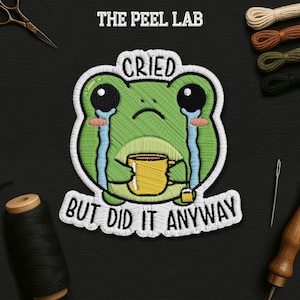 May include: Embroidered patch featuring a crying green frog holding a yellow mug. The patch has the text "Cried But Did It Anyway" and is surrounded by a white border. The Peel Lab is printed at the top.