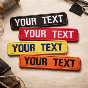 May include: Four rectangular embroidered patches in black, red, yellow, and orange, each with the text "YOUR TEXT" in white or blue. The patches are displayed on a beige fabric surface, with sewing tools visible.