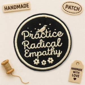 Peut inclure: Un écusson brodé noir avec les mots "Practice Radical Empathy" en écriture blanche, un oiseau et trois petites fleurs. L'écusson est entouré d'une bordure blanche. Il y a aussi des étiquettes en bois avec les mots "Handmade" et "Patch".