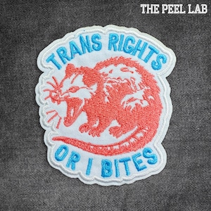 May include: Embroidered patch featuring a red opossum design and the text "TRANS RIGHTS OR I BITES" in blue. The patch has a white border and the text "THE PEEL LAB" in the top right corner.