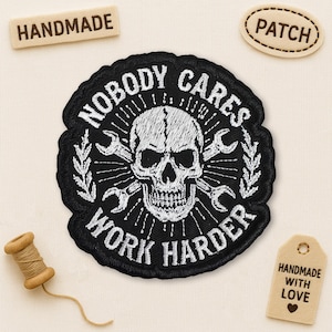 Peut inclure: Un écusson brodé noir et blanc avec un crâne et des clés à molette, entouré des mots "NOBODY CARES WORK HARDER". L'image comprend également de petites étiquettes avec les mots "HANDMADE" et "PATCH".