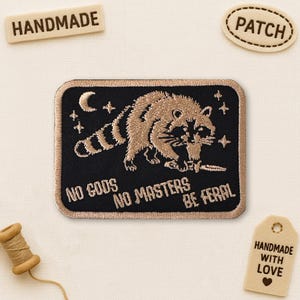 May include: Embroidered patch with a raccoon design, knife, stars, and crescent moon on a black background. Text reads "NO GODS NO MASTERS BE FERAL". Includes "PATCH", "HANDMADE" and "HANDMADE WITH LOVE" tags.