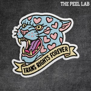 May include: Embroidered patch featuring a stylized jaguar head with a light blue base and pink heart-shaped spots. The jaguar has an open mouth with a purple tongue and the text "TRANS RIGHTS FOREVER" on a banner.