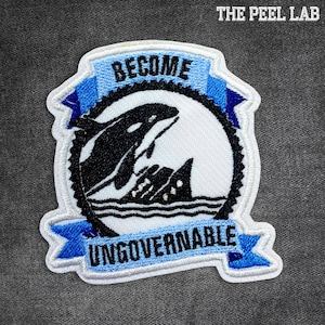 May include: Embroidered patch featuring a black and white orca breaching the water. The patch has a blue banner with the words "BECOME UNGOVERNABLE". The patch is bordered with a black and white stitched design. The text "THE PEEL LAB" is at the top.