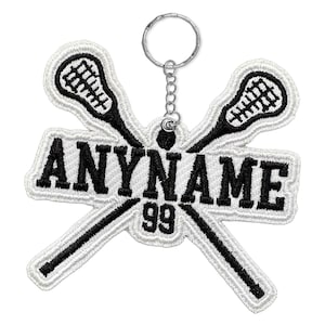 May include: A white and black embroidered lacrosse keychain. The design features two crossed lacrosse sticks with a net, and the text "ANYNAME 99" in bold black letters. A silver keyring is attached.