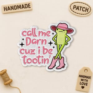 Peut inclure: Écusson brodé avec une grenouille verte portant un chapeau et des bottes roses, avec le texte "call me Darn cuz i be tootin'". Le patch a une bordure blanche et des étoiles noires.