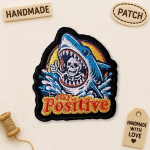 May include: Embroidered patch featuring a shark with a skeleton inside its mouth, with the words "Stay Positive" in red. The shark is blue and white, set against a yellow and black background. The patch is surrounded by a black border.