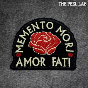 Peut inclure: Écusson noir avec les mots "MEMENTO MORI" et "AMOR FATI" en blanc. Une rose rouge est brodée au centre. "THE PEEL LAB" est imprimé en haut à droite.