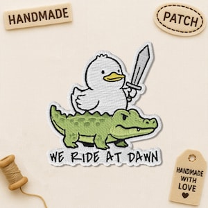 May include: Embroidered patch featuring a white duck holding a sword, riding a green alligator. The text "WE RIDE AT DAWN" is below the alligator. The word "PATCH" is on a separate patch.