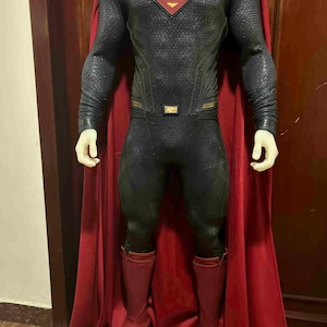 May include: A Superman costume featuring a black and gray textured bodysuit with a red and gold emblem on the chest. The costume includes a red cape and matching red boots. The overall design is a superhero theme.