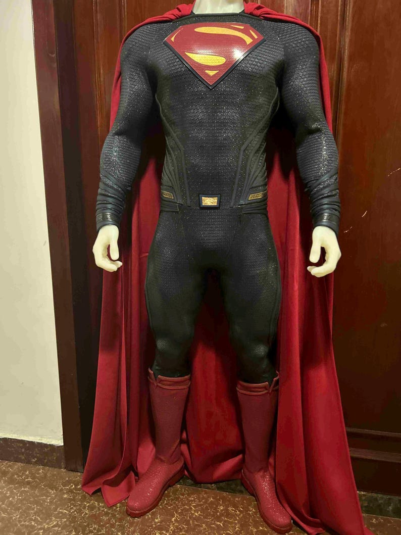 May include: A Superman costume featuring a black and gray textured bodysuit with a red and gold emblem on the chest. The costume includes a red cape and matching red boots. The overall design is a superhero theme.