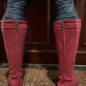 May include: A pair of knee-high, red boots with a textured pattern. The boots have a zipper running up the back and are paired with black, textured leggings. The boots are standing on a brown surface.