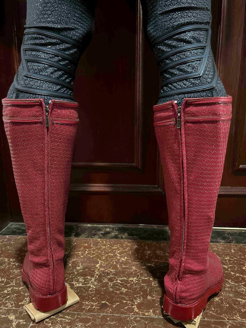 May include: A pair of knee-high, red boots with a textured pattern. The boots have a zipper running up the back and are paired with black, textured leggings. The boots are standing on a brown surface.